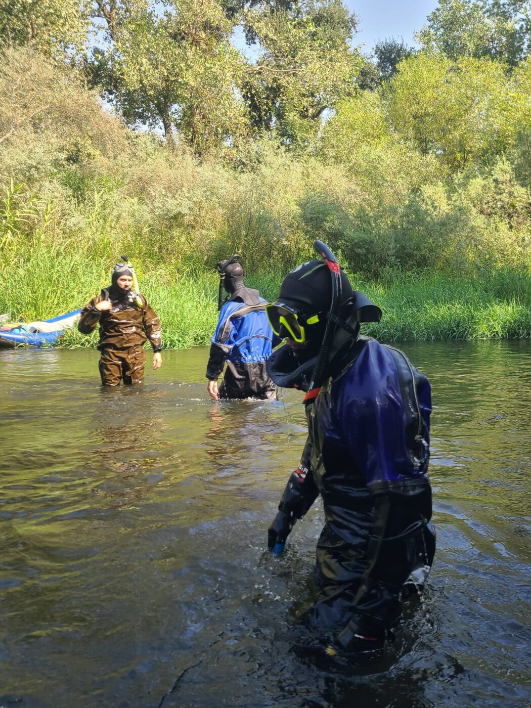 Three people in dry suits with snorkels on are standing in waist deep water surrounded by trees.